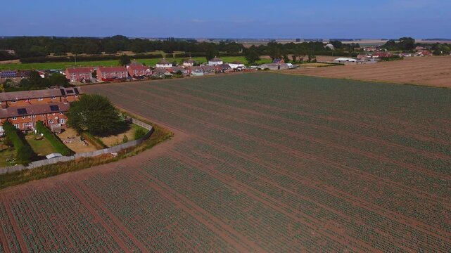 Aerial view of farmland beside village homes. Ridges stretch across rich, brown soil. Village rooftops peek through green trees. Sky glows blue and cloudless above