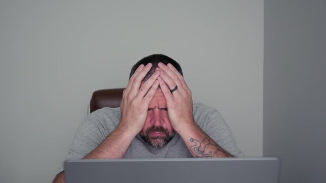 Stressed middle-aged man holding his head in pain while working on a laptop computer in a dimly lit home office, experiencing a migraine headache