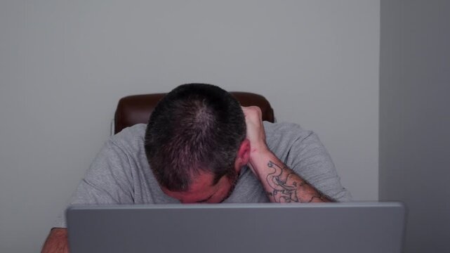 Stressed middle-aged man holding his head in pain while working on a laptop computer in a dimly lit home office, experiencing a migraine headache