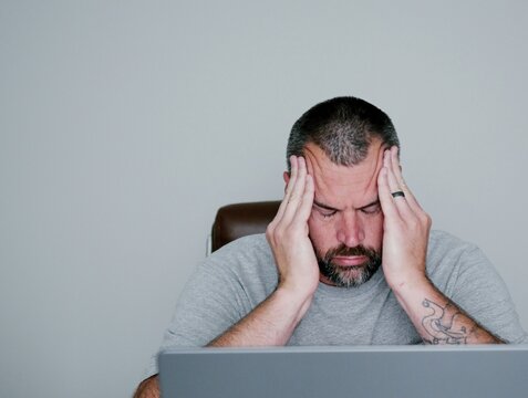 Stressed middle-aged man holding his head in pain while working on a laptop computer in a dimly lit home office, experiencing a migraine headache