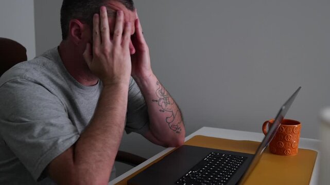 Stressed middle-aged man holding his head in pain while working on a laptop computer in a dimly lit home office, experiencing a migraine headache