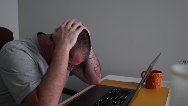 Stressed middle-aged man holding his head in pain while working on a laptop computer in a dimly lit home office, experiencing a migraine headache