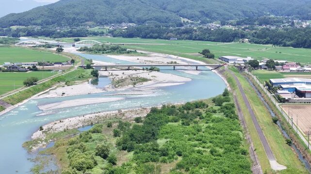 Aerial view captures a winding river flowing through green fields. Two bridges span the river, connecting land masses. Rail tracks run parallel to the river's edge