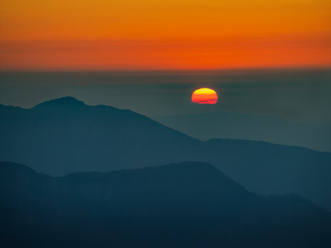 Telephoto landscape photograph looking east from the summit slopes of Grintovec (Grintavec) at dawn, capturing the glowing sun rising above the layered mountain silhouettes and the rocky crest