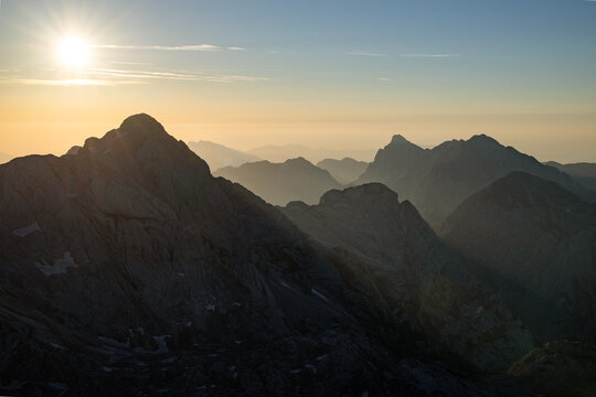 A magnificent alpine panorama shot east from the summit slopes of Grintovec (Grintavec) at sunrise, featuring a brilliant sunburst over the massive peak of Skuta with Kranjska Rinka and Mrzla Gora