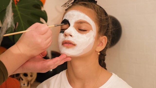 A cute teenage girl is getting ready for Halloween and doing face painting, with a skeleton painted on her face. October is a time of horror.