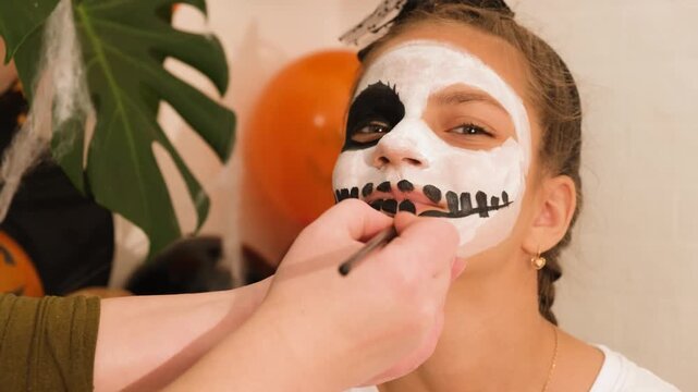A cute teenage girl is getting ready for Halloween and doing face painting, with a skeleton painted on her face. October is a time of horror.