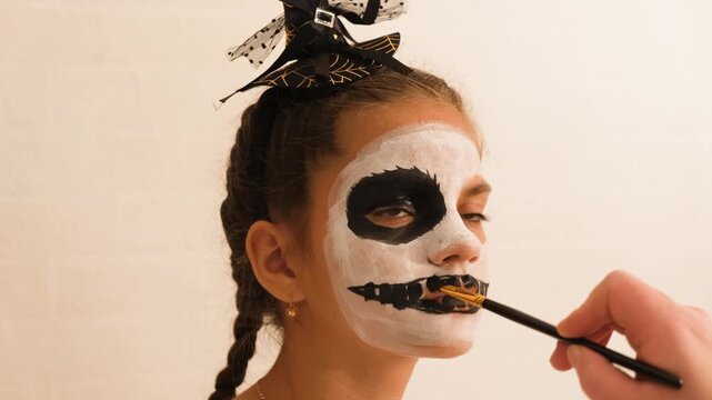 A cute teenage girl is getting ready for Halloween and doing face painting, with a skeleton painted on her face. October is a time of horror.
