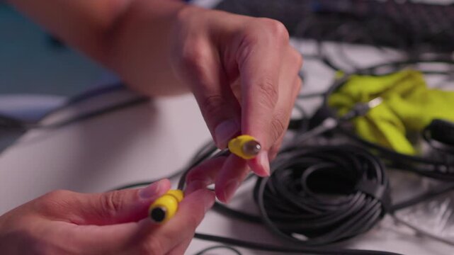 Hands aligning yellow electrical connector halves on black cable over tabletop workspace, showing careful joining motion, exposed pin, coiled wire, green fabric backdrop