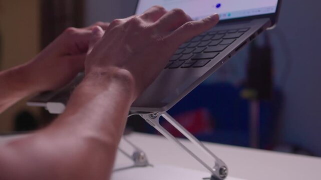 Hands typing on laptop raised by foldable metal stand on white desk, showing ergonomic workstation setup, active keyboard use, slight forward tilt, compact modern office scene