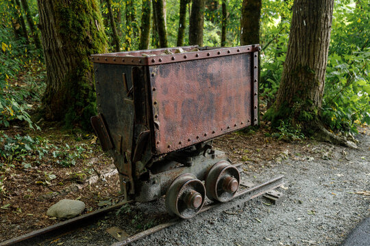 A rusted riveted mine ore cart loaded with rock sits on a short length of rail track in a green forest at Lafarge Lake park in Coquitlam, a nod to the area's mining heritage.
