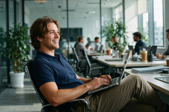 Young man in office smiling and relaxing in sleek chair by open laptop with bright meeting rooms and coworkers behind him.