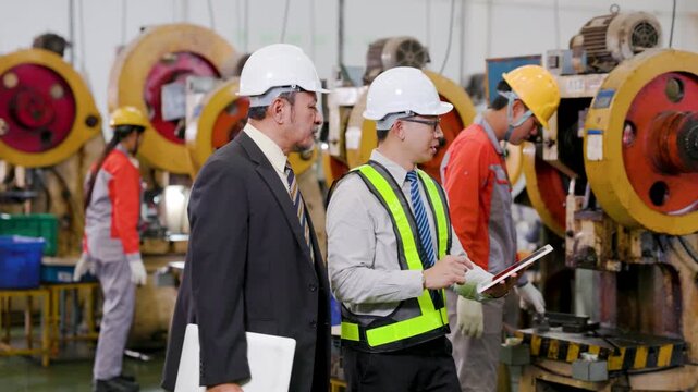 Engineers in hard hats review data on a tablet inside a manufacturing plant, discussing operations amid machinery while workers handle equipment in the background