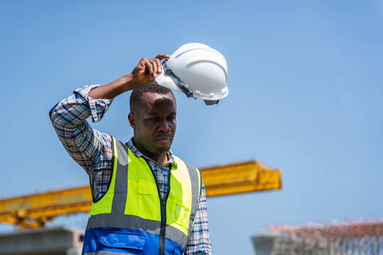 Exhausted engineer taking a break at construction site in heat, Construction worker holding hard hat at infrastructure project site, Portrait of worker man with hard hat against blue sky