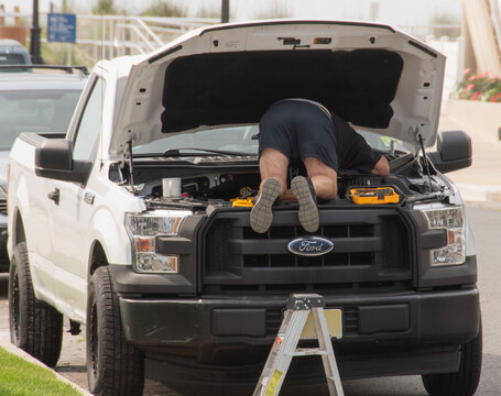 A mechanic leans over the open hood of a white Ford pickup truck parked on a residential street, using tools and a ladder to inspect and repair the engine and front components