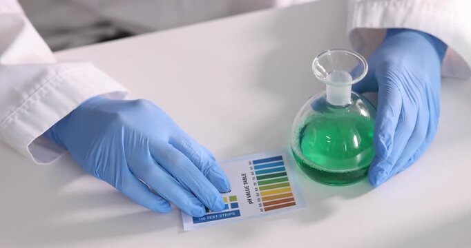 Scientist holding flask with liquid and color table for PH testing at white table in laboratory, closeup