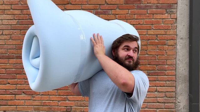 4K video. Young adult man with beard stands near brick wall of house and holds blue bundle of foam rubber for sofa, looks back. Concept of furniture repair, delivery, rent apartment, dream, sleep