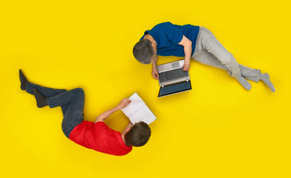 Two relaxed people lying on bright yellow floor or background seen from above, one using a laptop, one reading a book