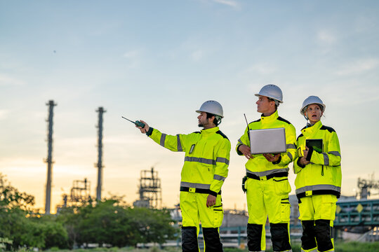 Three petrochemical workers in yellow hi vis suits and white hard hats as an outstretched arm directs attention to the far storage area on an open refinery yard at nightfall