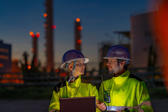 Young male oil and gas engineer in a yellow hi vis jacket and white helmet looking over his shoulder toward a sound in the dark on night watch before an illuminated refinery skyline