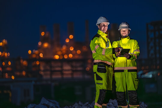 Two male oil and gas engineers in yellow hi vis suits and white hard hats as the two agree on the next stop of the night patrol consulting a glowing tablet at a night time refinery