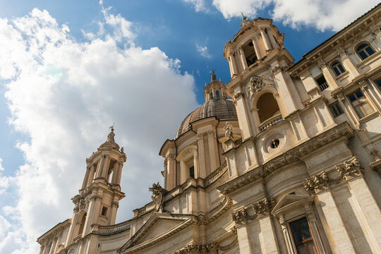 Vista contrapicada de la iglesia barroca de Sant'Agnese in Agone en Roma. Su imponente c&uacute;pula y campanarios se alzan contra un dram&aacute;tico cielo azul con nubes blancas.