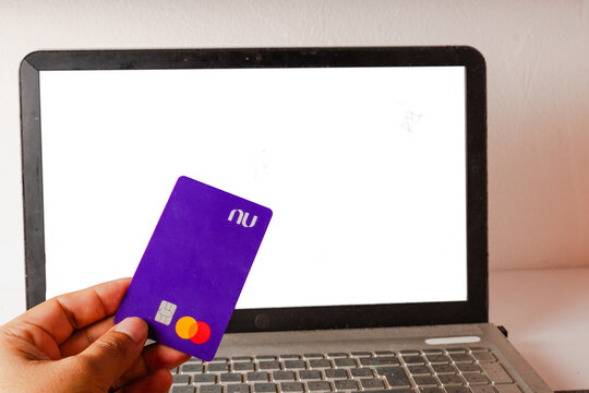 Hand holding purple Nubank credit card vertically in front of open laptop screen closeup