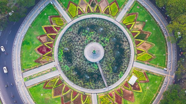 Sunrise illuminates MalangGolden hour bathes historic building and central monument. Circular pond reflects sky and structure. Lush greenery surrounds the scene. Distant trees frame the peaceful vista