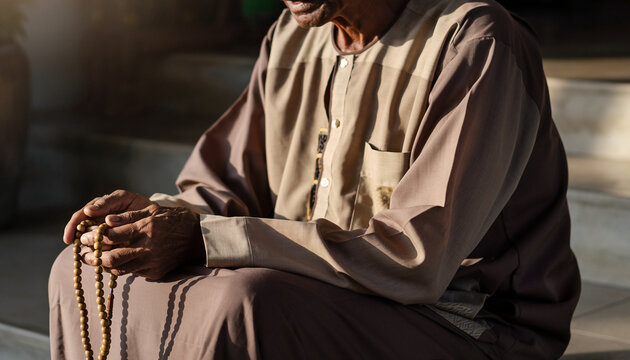 An elderly person sitting outdoors, holding prayer beads, wearing traditional clothing, with a peaceful expression, and a blurred background suggesting a calm, contemplative moment.