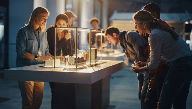 People gathered around an exhibition display case, observing architectural models or artifacts, with warm lighting highlighting the scene outdoors during evening hours.