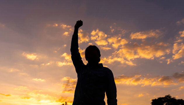 Silhouette of a person raising their arm against a vibrant sunset sky with scattered clouds, creating a dramatic and inspiring scene.