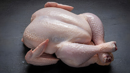 Naklejka premium Whole Raw Chicken Ready for Cooking: A whole, uncooked chicken lies on a dark, textured surface, prepared for roasting or other culinary preparations.
