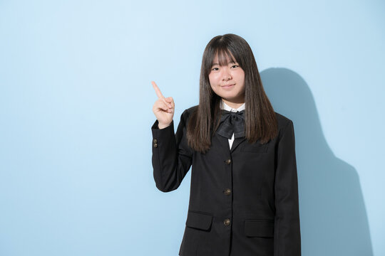 水色背景で制服で立つカメラ目線笑顔の女子中学生や高校生のイメージ　指差しでポイントのポーズ