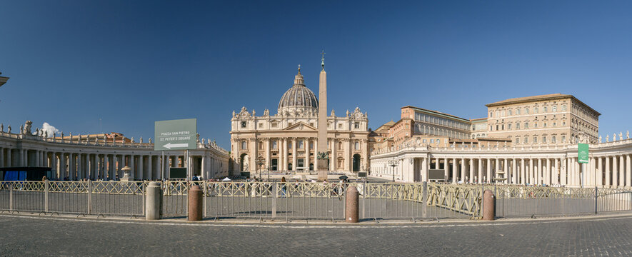 Vista panor&aacute;mica de la Plaza de San Pedro y la Bas&iacute;lica en el Vaticano, Roma. D&iacute;a soleado con cielo azul, obelisco y columnatas de Bernini. Algunos turistas y vallas visibles.