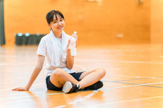 Elementary, middle, and high school girls in tracksuits rehydrating with bottled water in the gymnasium.