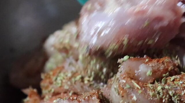Raw chicken pieces being seasoned with various dried herbs and spices in stainless steel bowl for cooking preparation.