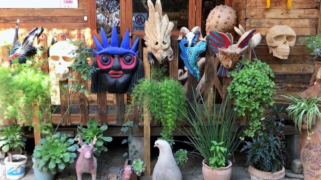 Front view of decorative handcrafted masks, sculpted ornaments, and potted plants displayed outside a rustic artisan shop creating a colorful cultural storefront during daytime