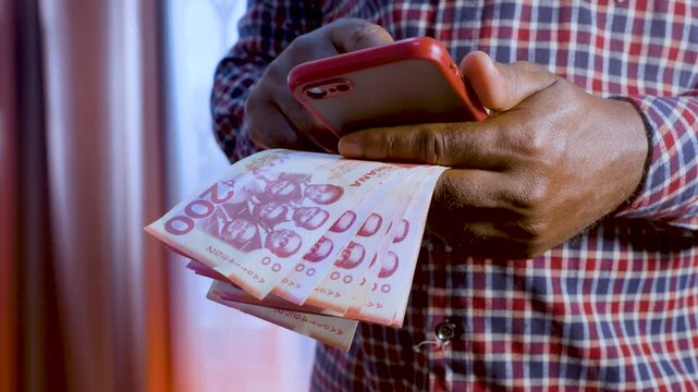 A close-up shot of a man wearing a checkered shirt, holding a stack of 200 Ghanaian cedi banknotes in one hand while typing or navigating on a red smartphone with the other, symbolizing mobile banking
