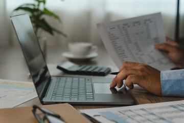 Fototapeta premium Business person reviews printed financial records while using a laptop and calculator at a desk, checking figures and documents for accounting analysis and expense report preparation.