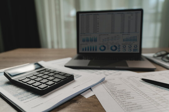 Calculator on printed financial reports beside laptop dashboard and pen on desk, showing accounting document review, budgeting workflow, and business data analysis for reporting tasks.
