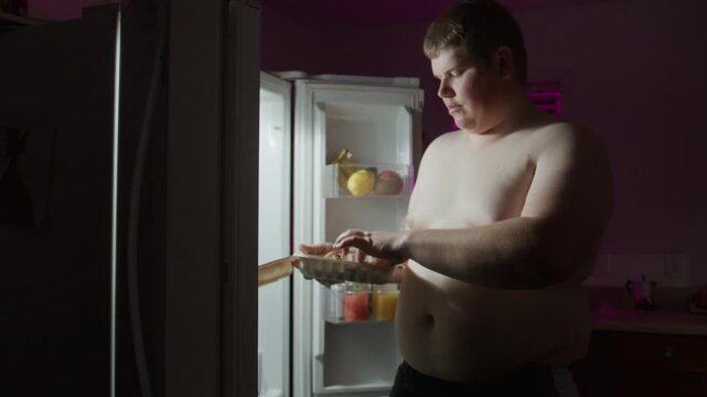 Overweight Man Eating Late at Night from Open Refrigerator. Overweight man eating a night snack near refrigerator. Emotional eating, unhealthy lifestyle, obesity, overeating, binge eating at night.