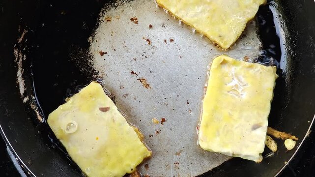Close up view of three rectangular pieces of tempeh coated in yellow batter being fried in a black pan with bubbling hot oil