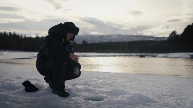 Bundled man crouches on river ice, jigging with a short fishing rod over a small hole as low sun reflects off the water.