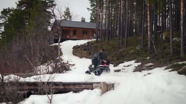 A snowmobile rider drives up a slushy track toward a remote wooden cabin in the forest. Ideal for travel, transport, cabin life and outdoor adventure content.