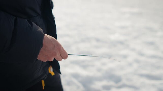Tight side view of a bundled person slowly jigging a short rod over bright ice on a frozen lake surface.
