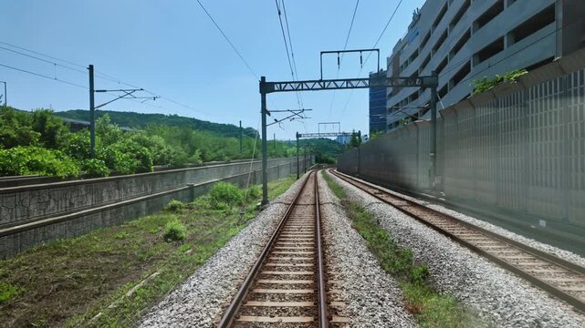 A front-facing view from a train showing railway tracks leading through an open area with overhead electric lines and green hills.