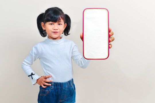 Cheerful little Asian girl with pigtails hair smiling excitedly while showing a blank smartphone mockup screen against an isolated clean background.
