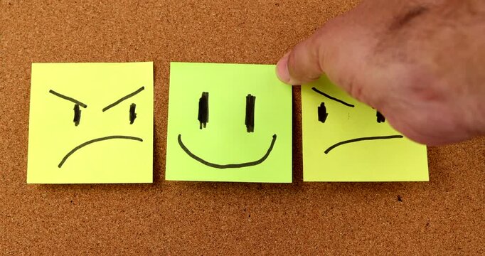A person hand placing a bright fluorescent yellow happy face sticky memo note in the middle of multiple blue sad frowning paper notes on a brown office corkboard
