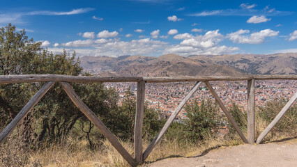 Fototapeta premium High-altitude observation deck. Behind the wooden railings, you can see the city from a height. Lots of buildings with red tiled roofs. Mountains against a blue sky and clouds. Peru. Cusco.