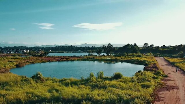 Aerial drone footage of a tranquil lakeside landscape surrounded by lush green vegetation and residential area under a clear blue sky.